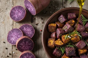 A close-up shot of a smooth, vibrant lavender-colored purple sweet potato mash in a white ceramic bowl, garnished with a decorative swirl of white coconut cream and a small sprig of fresh green herbs on a rustic wooden background.